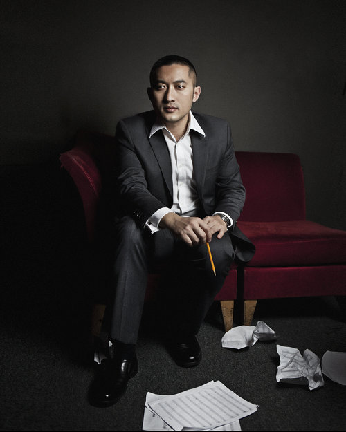 Chinese born composer Huang Ruo is seated on a red couch in the darkend room, holding a yellow pencil, as sheets of music, with some pages crumpled, lay at his feet.