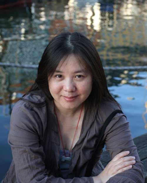 Headshot of Chinese-American composer Kui Dong seated on a wooden bench with watery reflections behind her in the background.