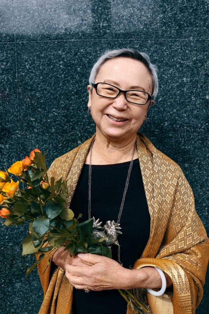 Chinese-American poet Flo Oy Wong holds a small bouquet of yellow and orange roses, as she stands in front of the shiny dark gray granite wall after The Last Hoisan Poets perform with Del Sol Quartet in Union Square, San Francisco. 