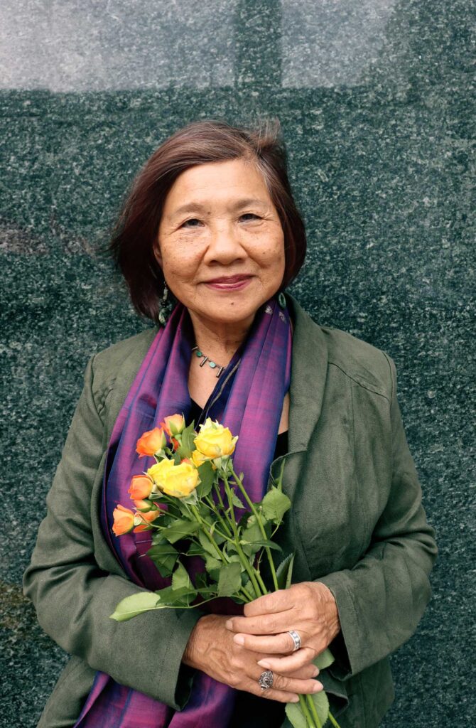 Chinese-American poet Genny Lim smiles, holding a small bouquet of yellow and orange roses, as she stands in front of the shiny dark gray granite wall after The Last Hoisan Poets perform with Del Sol Quartet in Union Square, San Francisco. 