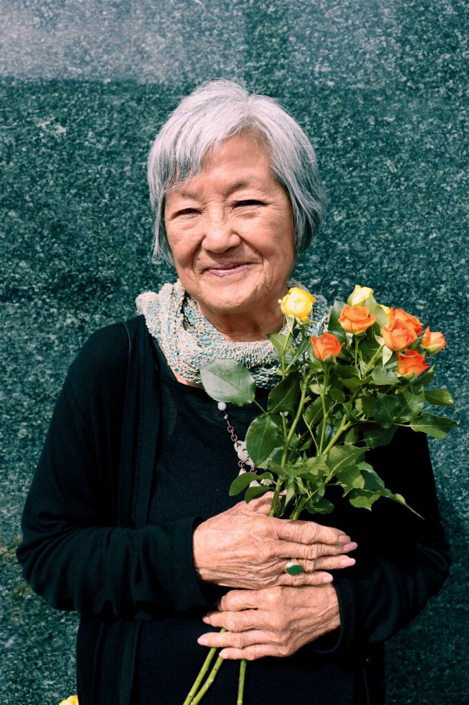 Chinese-American poet Nellie Wong smiles, holding a small bouquet of yellow and orange roses, as she stands in front of the shiny dark gray granite wall after The Last Hoisan Poets perform with Del Sol Quartet in Union Square, San Francisco. 